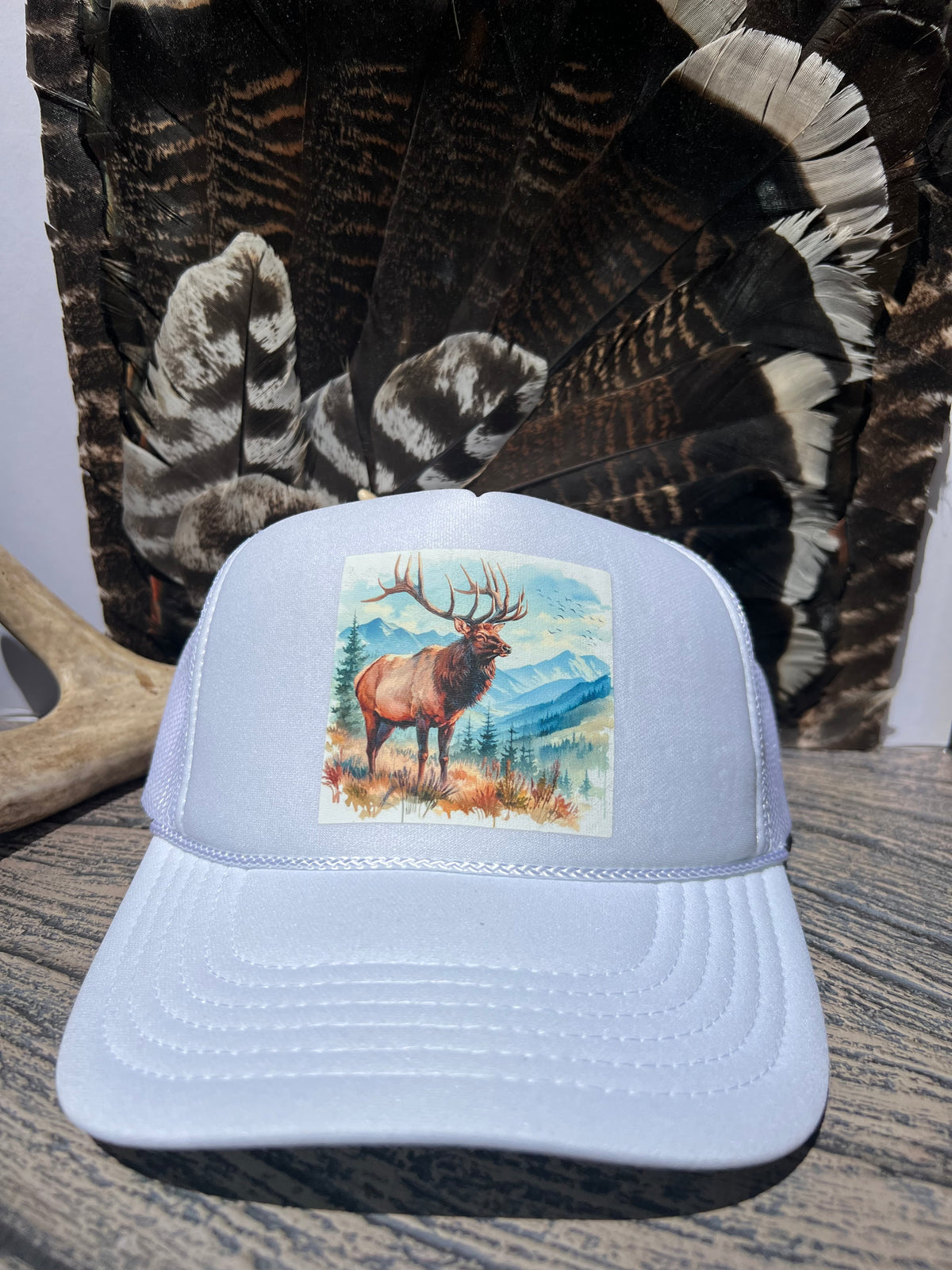A light blue trucker hat with a square front patch showing an elk standing in a mountain landscape, displayed on a wooden surface with a large feather bundle in the background.