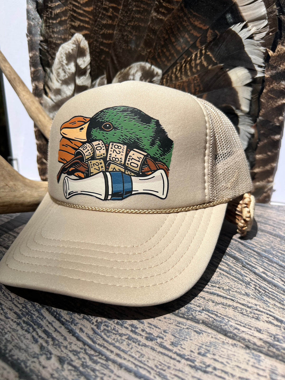 A beige trucker hat with a green and orange duck graphic on the front, styled on a rustic wooden surface with deer antlers and feathers in the background.