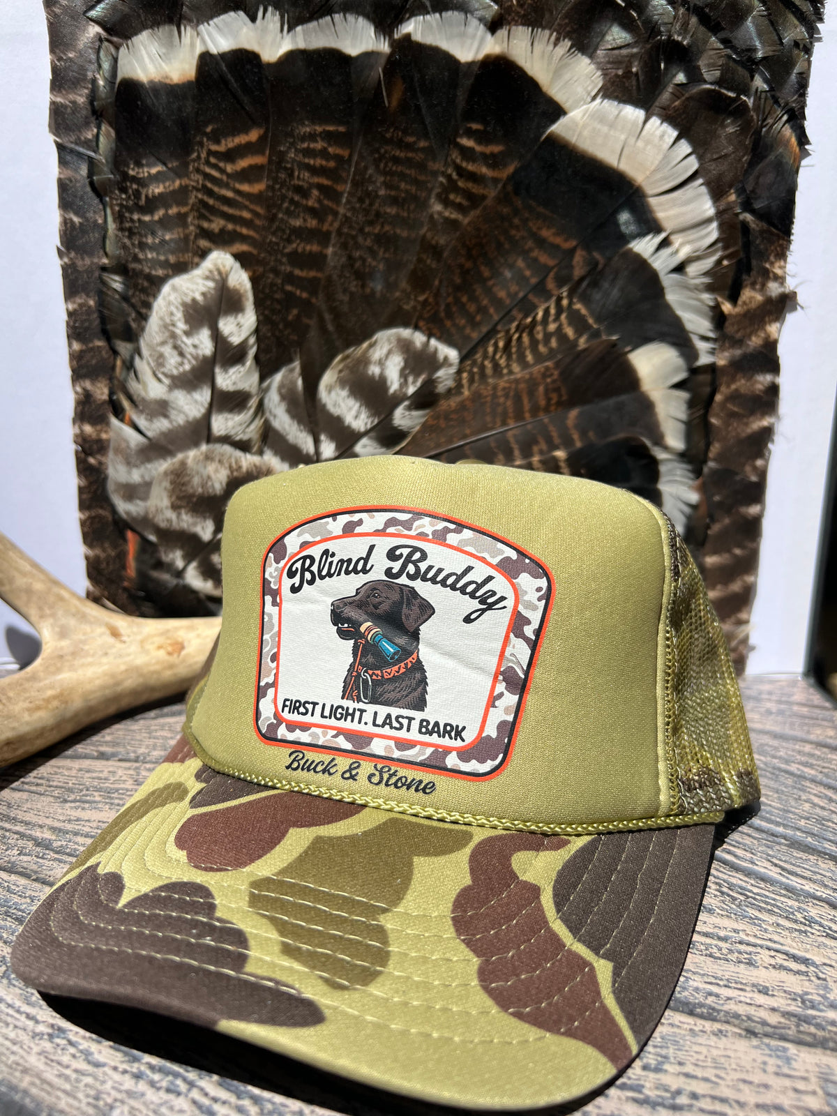 A camouflage trucker hat with a front patch reading 'Blind Buddy' and 'Buck & Stone' is displayed on a wooden surface in front of a large turkey tail backdrop.