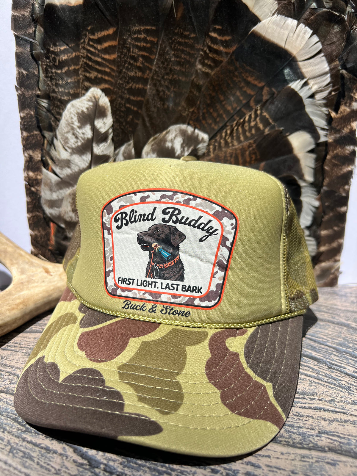 A camouflage trucker hat with an olive green crown and a curved brim featuring a brown and olive camo pattern. The front panel has a patch reading 'Blind Buddy' with a black dog illustration and 'Buck & Stone' below it.