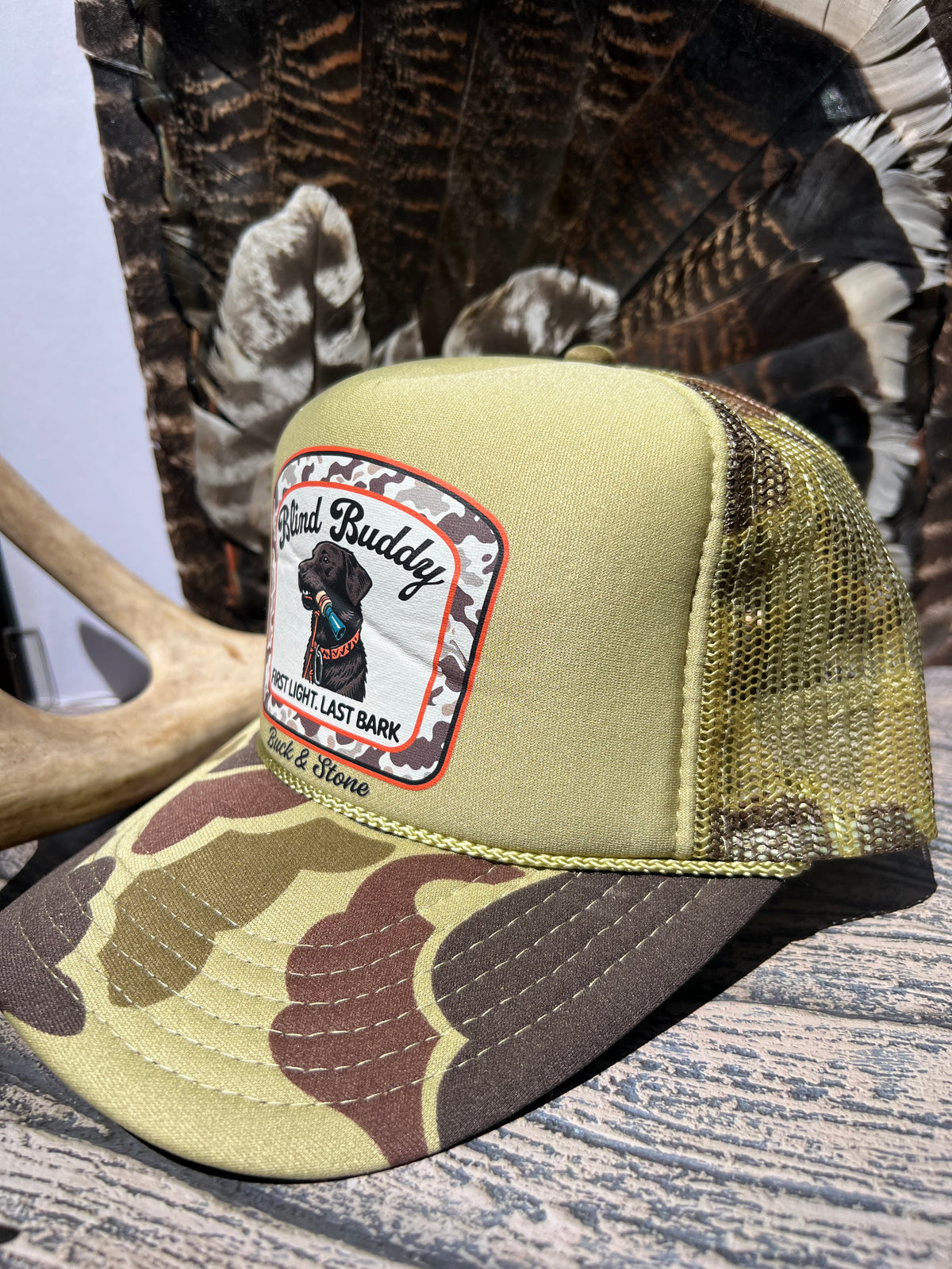 A camouflage trucker hat with a tan mesh back, featuring a front patch that reads 'Fired Up Buddy' and 'You Are My Last Bark' with a black dog illustration. The hat is styled on a rustic wood surface with a deer antler and a large feather fan in the background.
