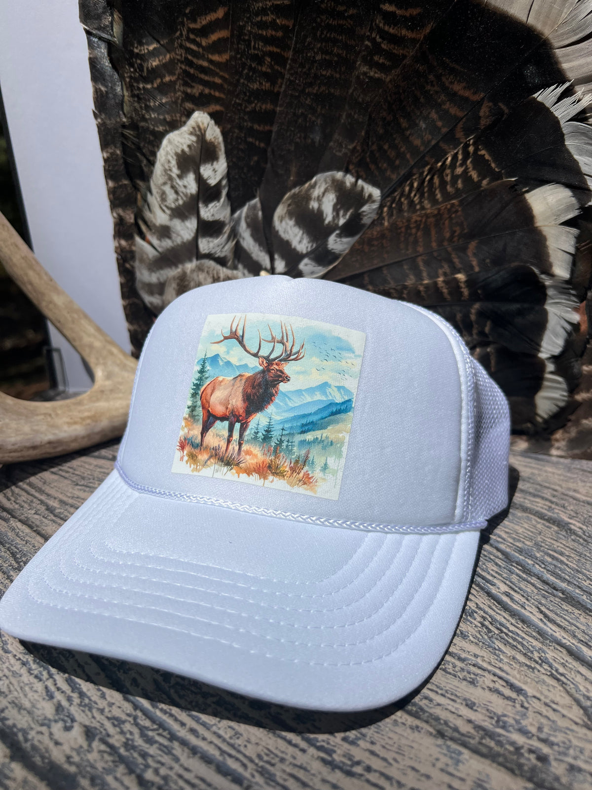 A white trucker hat with a square front patch showing a painted elk in a mountain landscape, displayed on a rustic wooden surface with feathers and antlers in the background.