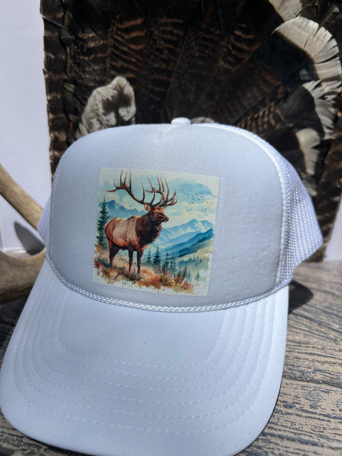 A white trucker hat with a printed graphic of an elk in a mountain landscape on the front panel, displayed on a rustic wooden surface with camouflage and deer antlers in the background.