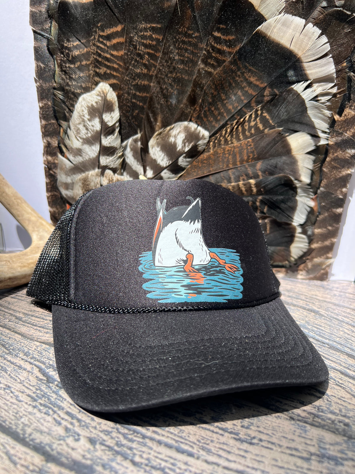 A black trucker hat with a graphic of a duck diving into blue water, displayed on a wooden surface with a large feather bundle in the background.