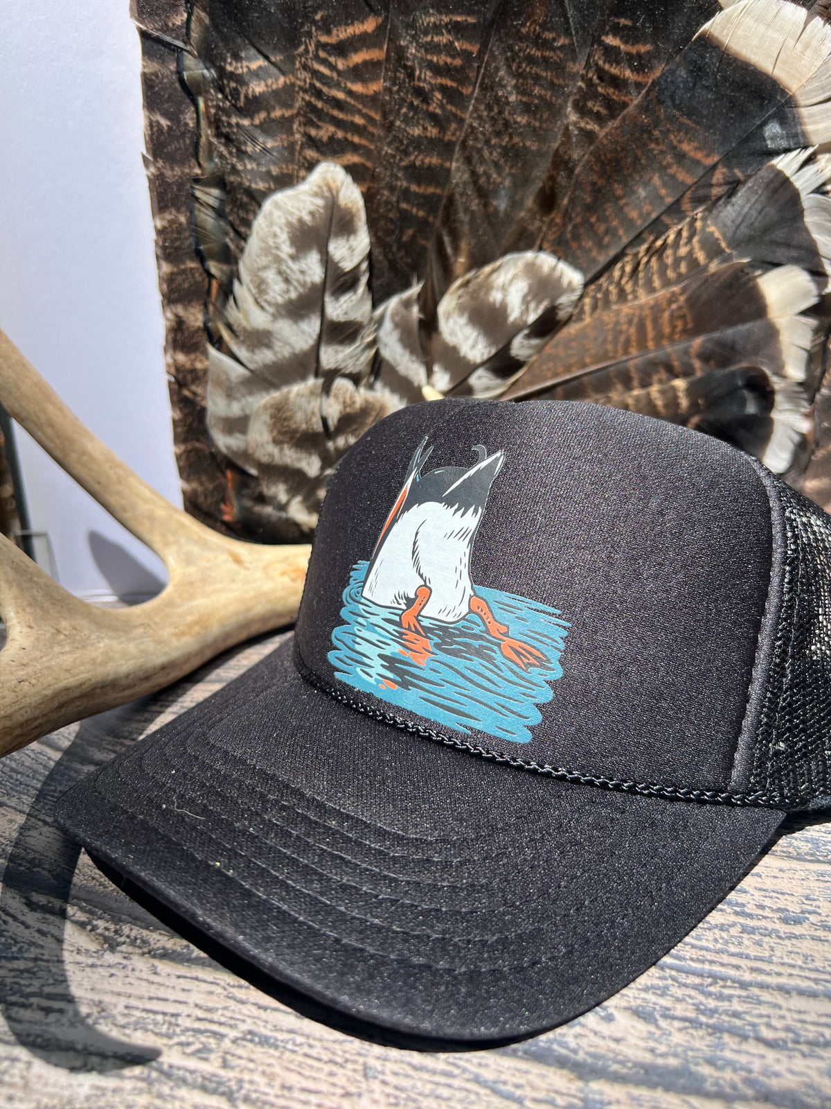 A black trucker hat with a graphic of a duck hunting in blue water, styled on a wooden surface with a deer antler and wild turkey feathers in the background.