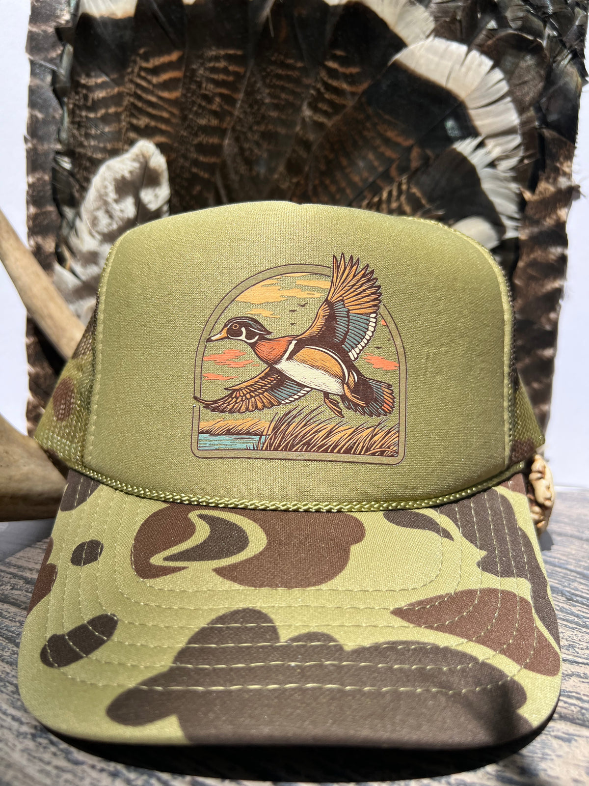 An olive green camouflage trucker hat with a detailed embroidered duck illustration on the front panel, displayed against a rustic camouflage and wood background.