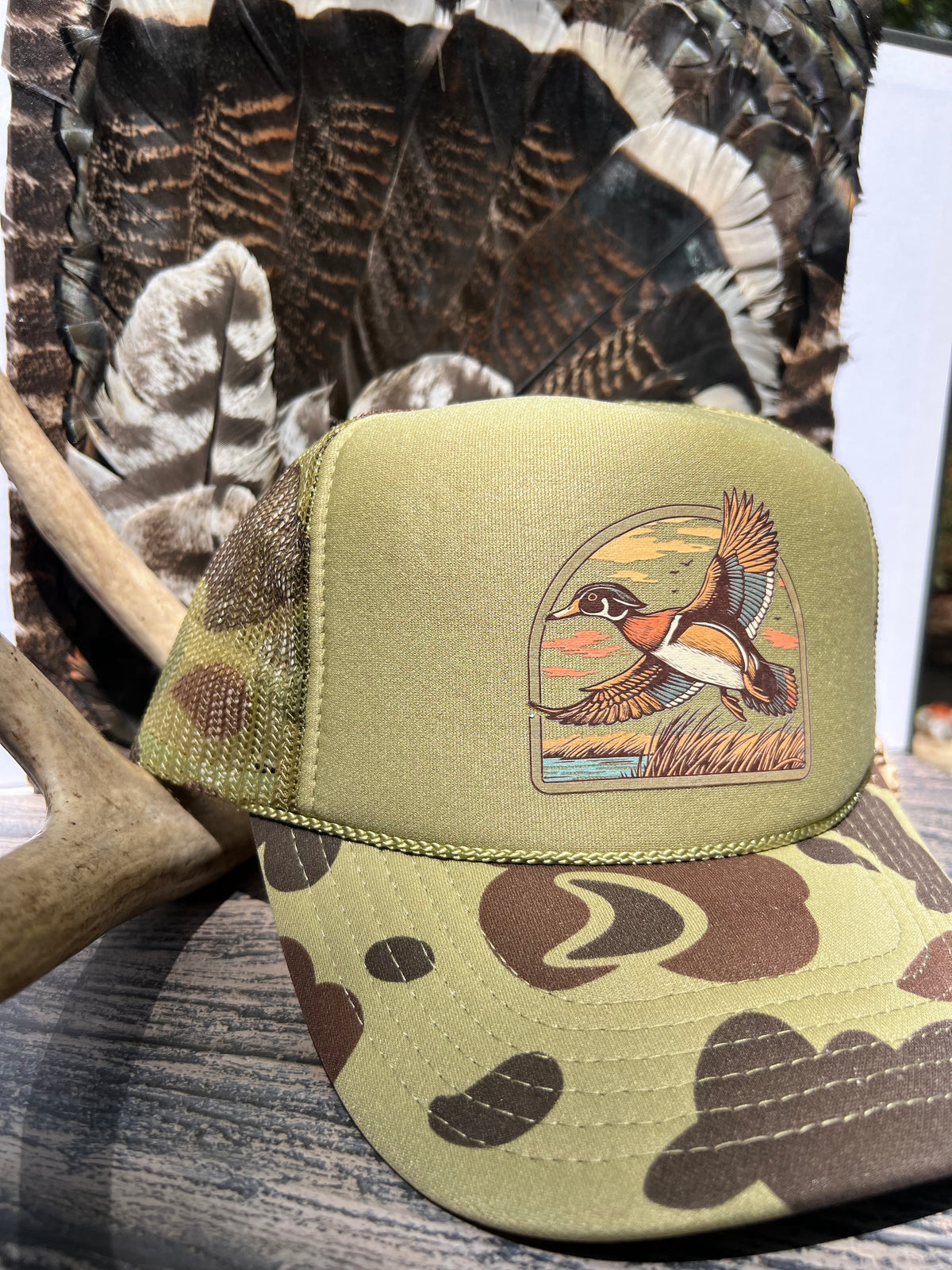 A camouflage trucker hat with an embroidered wood duck illustration on the front panel, styled with a deer antler and camouflage feathers in the background.