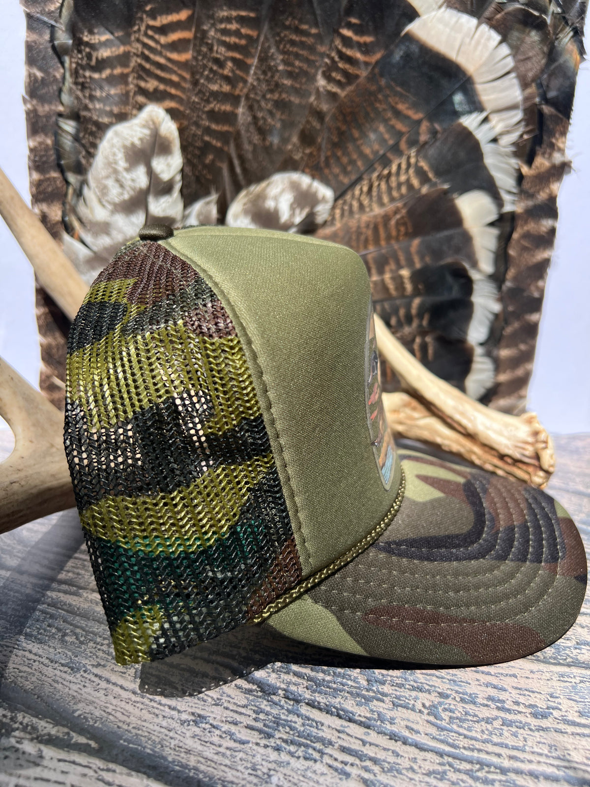 An olive green camouflage trucker hat with a mesh back, styled on a rustic wooden surface with a deer antler and a large turkey feather backdrop.