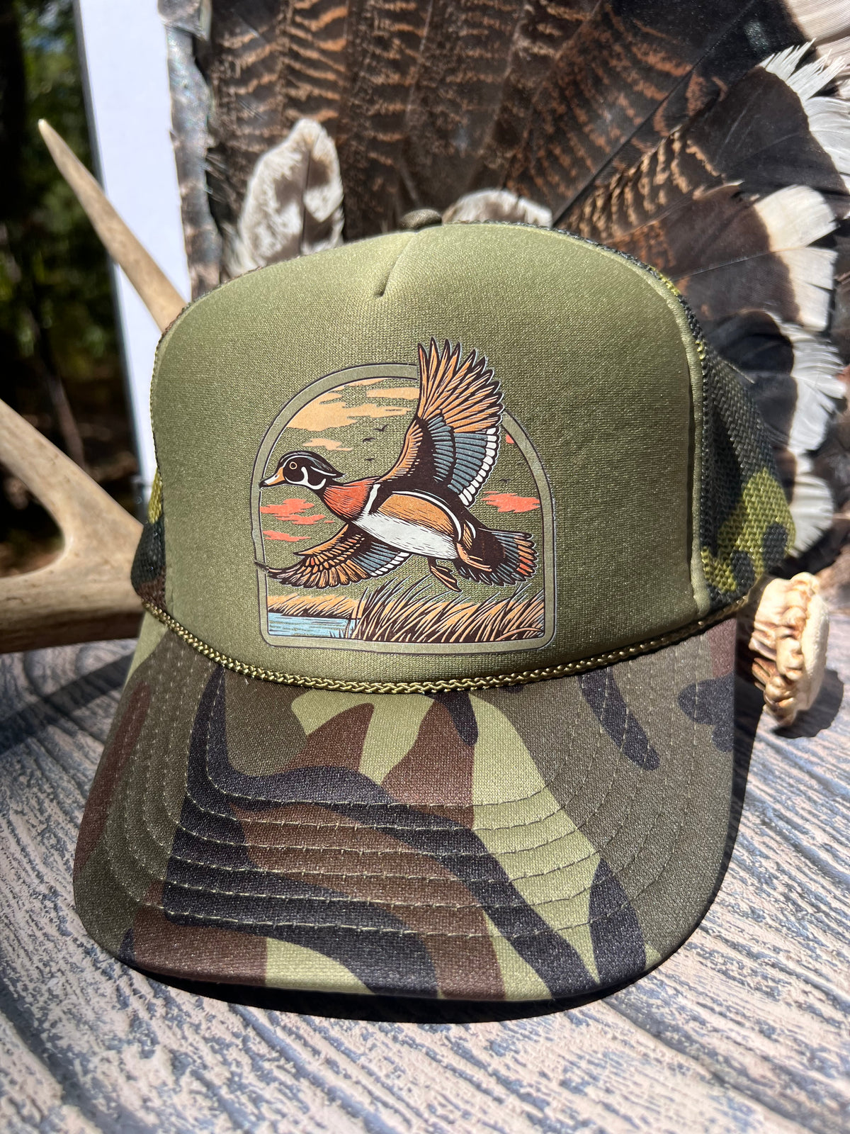 An olive green camouflage trucker hat with a detailed embroidered chest graphic of a flying duck, displayed on a rustic wooden surface with antlers and feathers in the background.