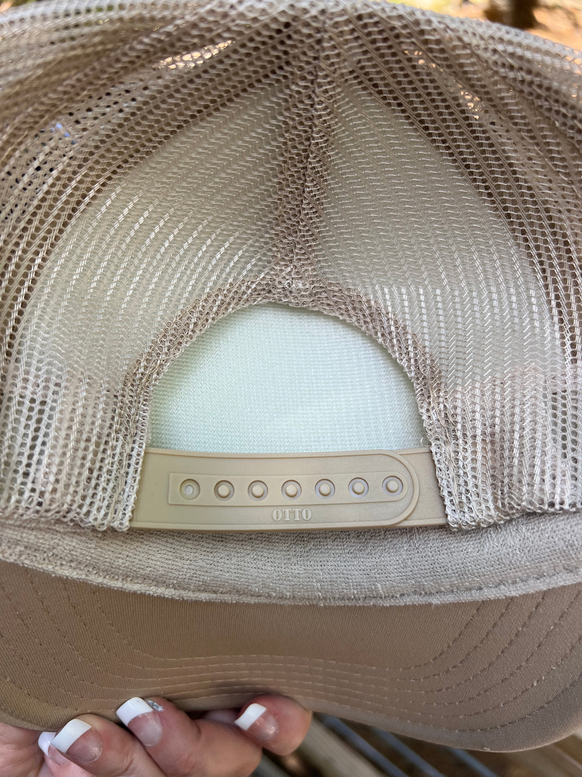 Close-up of the back of a beige trucker hat with a tan mesh back and a beige plastic snap closure. A hand with white manicured nails is holding the brim at the bottom.