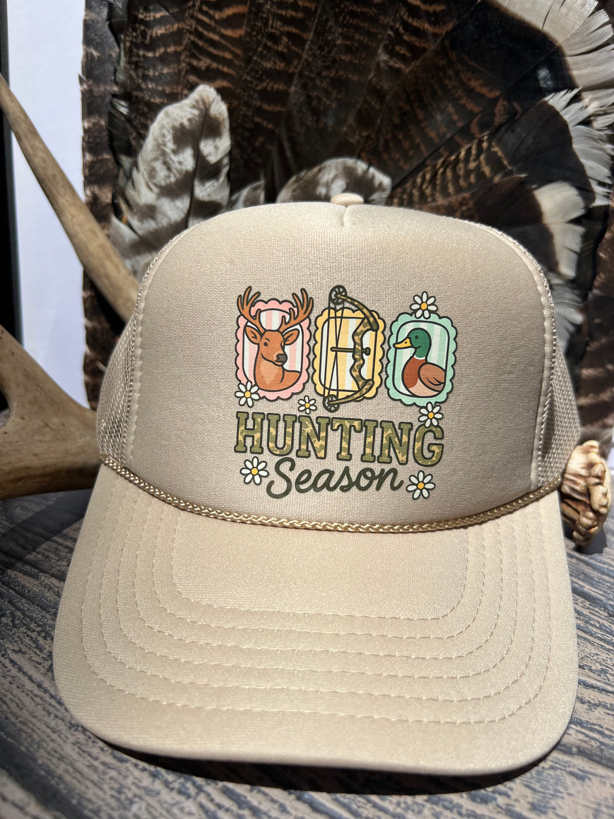 A beige trucker cap with a 'Hunting Season' graphic on the front, featuring deer, a bow, and duck illustrations, displayed on a rustic wood surface with antlers and feathers in the background.