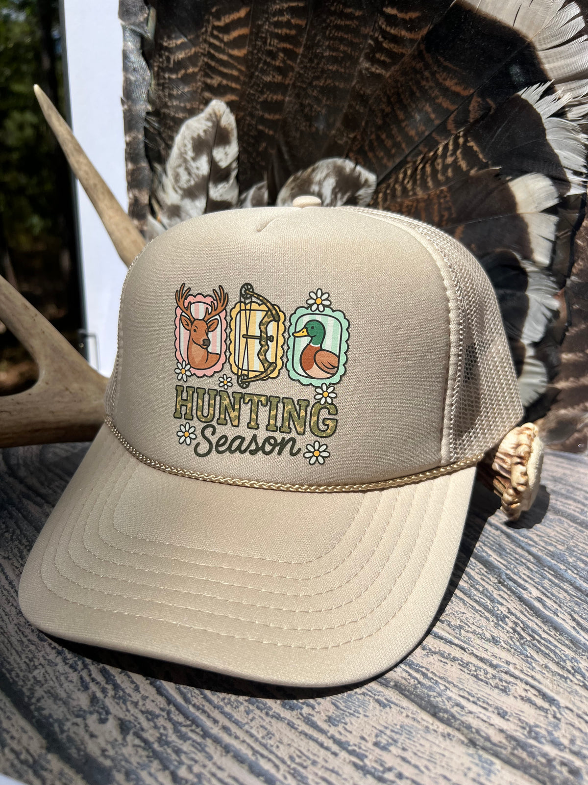 A beige trucker hat with a colorful embroidered 'HUNTING Season' graphic featuring a deer, turkey, and duck, styled on a rustic wood surface with antlers and wild turkey feathers in the background.