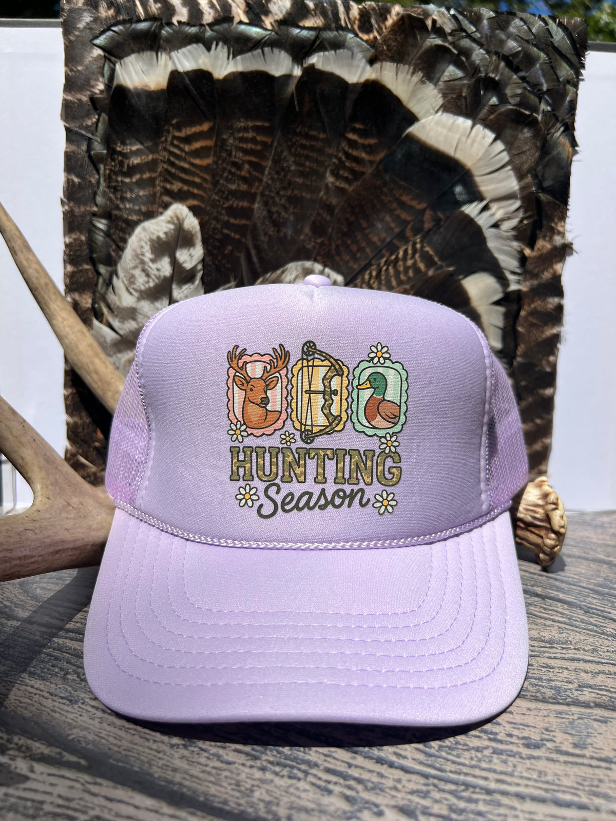 A lavender trucker hat with an embroidered 'Hunting Season' graphic featuring deer, a turkey, and snowflakes, displayed on a wooden surface with a large turkey feather and antler in the background.