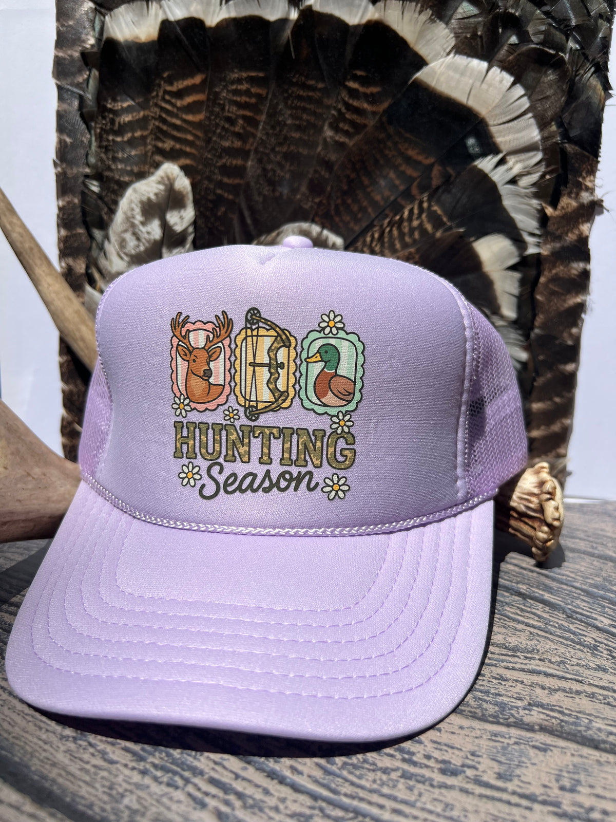 A lavender purple trucker hat with a 'Hunting Season' graphic on the front, styled on a rustic wooden surface with a large feather bundle in the background.