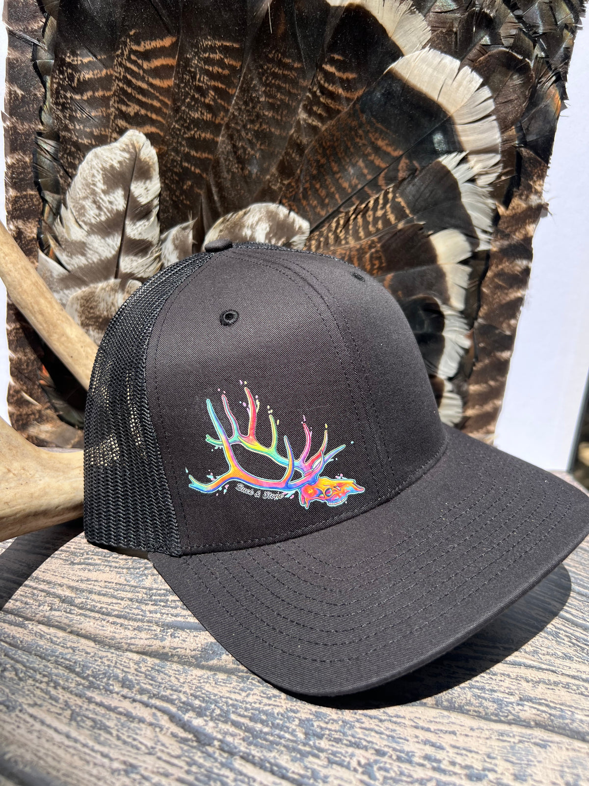 A black trucker hat with a colorful embroidered antler design and the words 'Wild Turkey' on the front, displayed on a rustic wooden surface with a feathered chair back in the background.