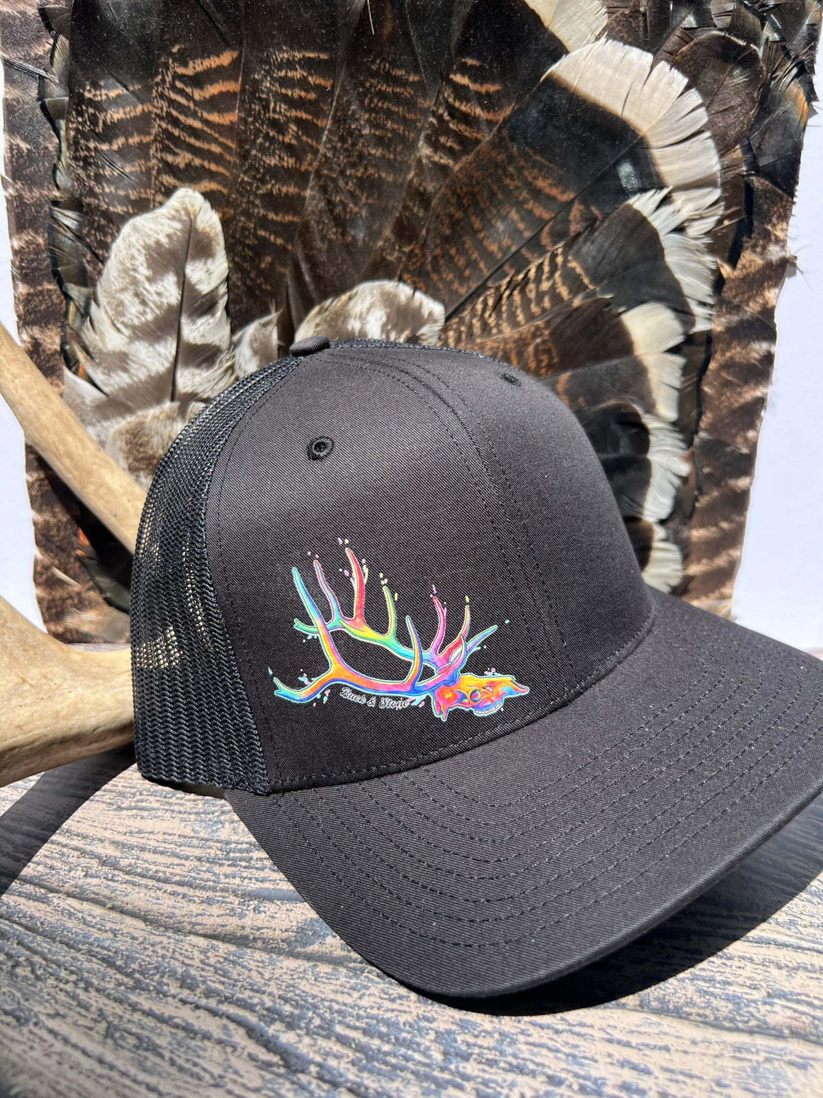 A black trucker-style baseball cap with a colorful embroidered deer antler and fish design on the front, displayed outdoors against a background of large feathers and wood.