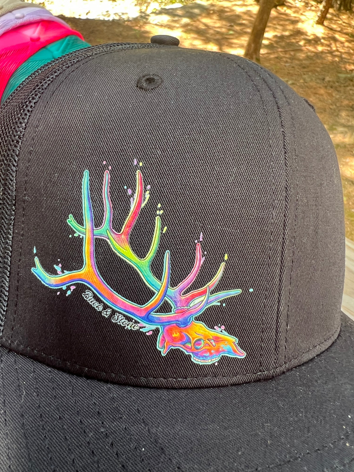 A close-up of a black trucker cap featuring a vibrant, multicolored antler graphic on the front panel. The graphic has a paint-splatter effect with rainbow hues including green, yellow, orange, pink, and blue.