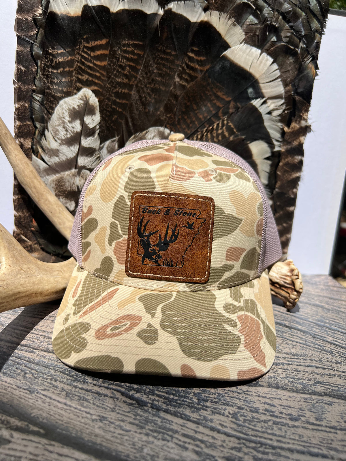 A camouflage trucker hat with a brown leather patch on the front that reads 'Duck & Deer Hunting Club' with a deer antler graphic, displayed on a wooden surface with a large turkey feather backdrop.
