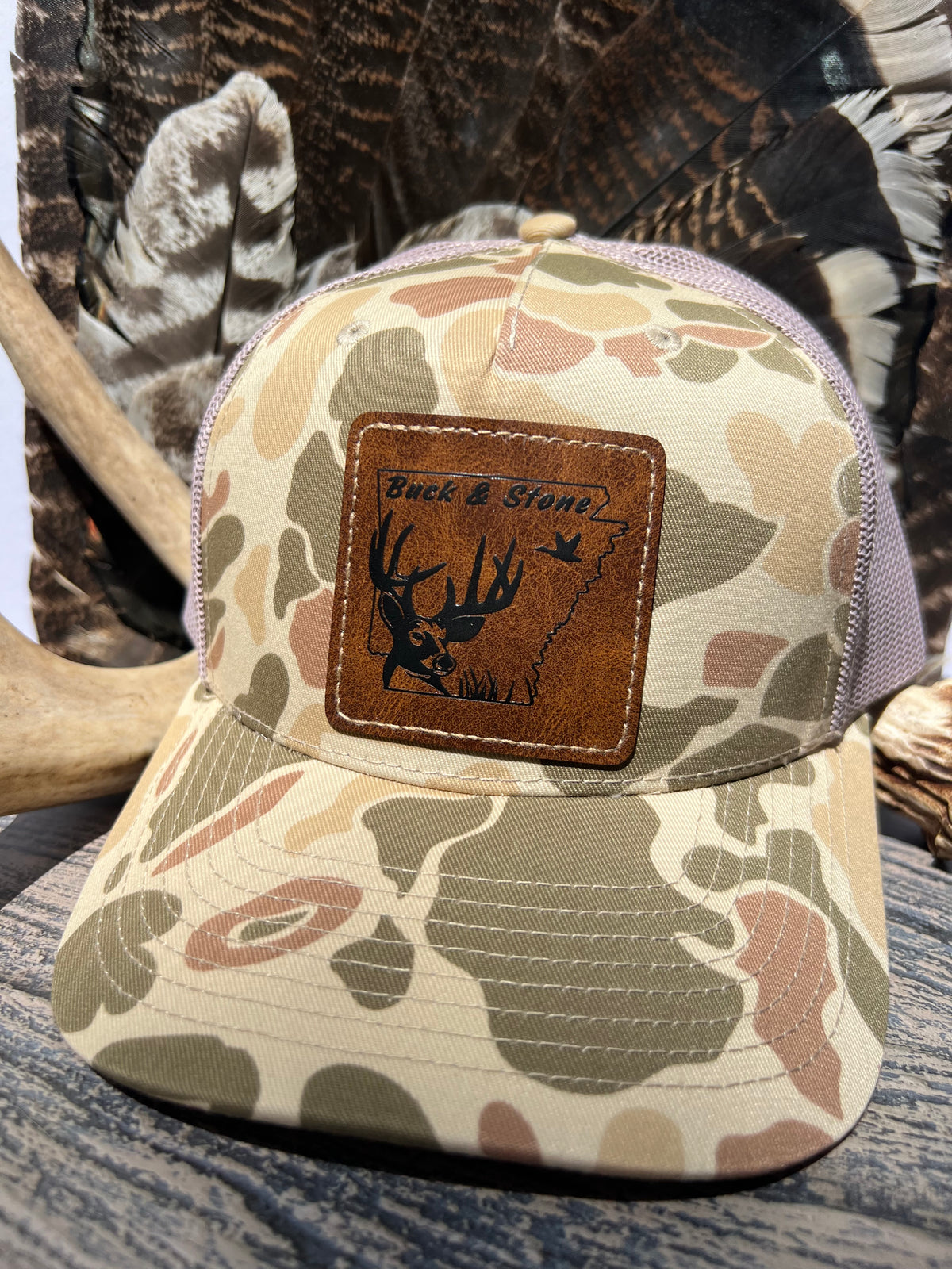 A camouflage trucker hat with a brown leather patch on the front featuring a deer graphic and the text 'Buck & Stone'. The hat is styled on a rustic wood surface with antlers and a camouflage-patterned background.