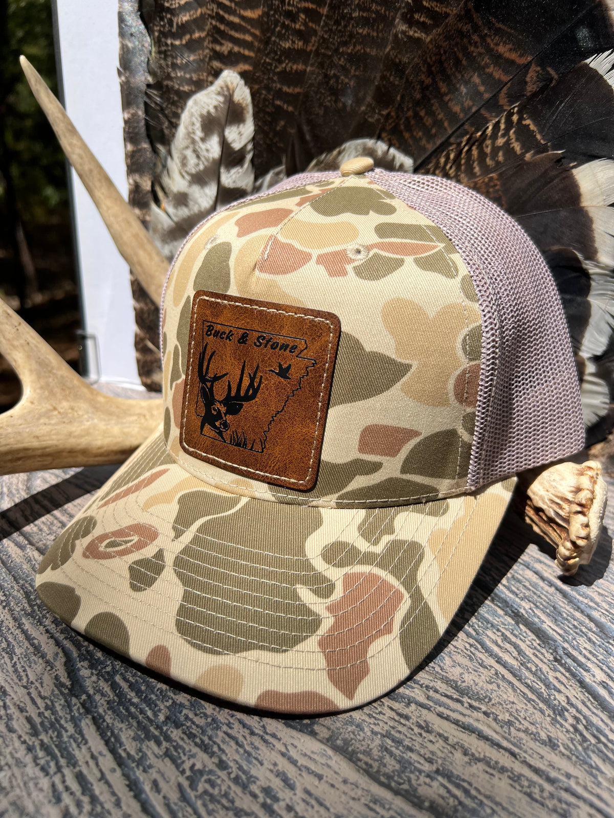 A camouflage trucker hat with a brown leather patch on the front that reads 'Back & Shot' with a deer graphic, displayed on a rustic wood surface with antlers and feathers in the background.