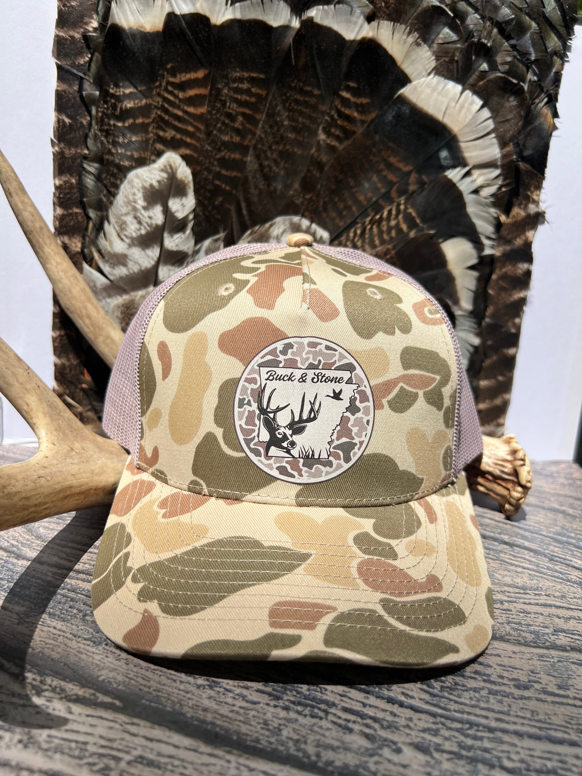 A camouflage trucker hat with a circular 'Buck & Bone' logo patch on the front, styled on a wooden surface with deer antlers and a large turkey feather backdrop.