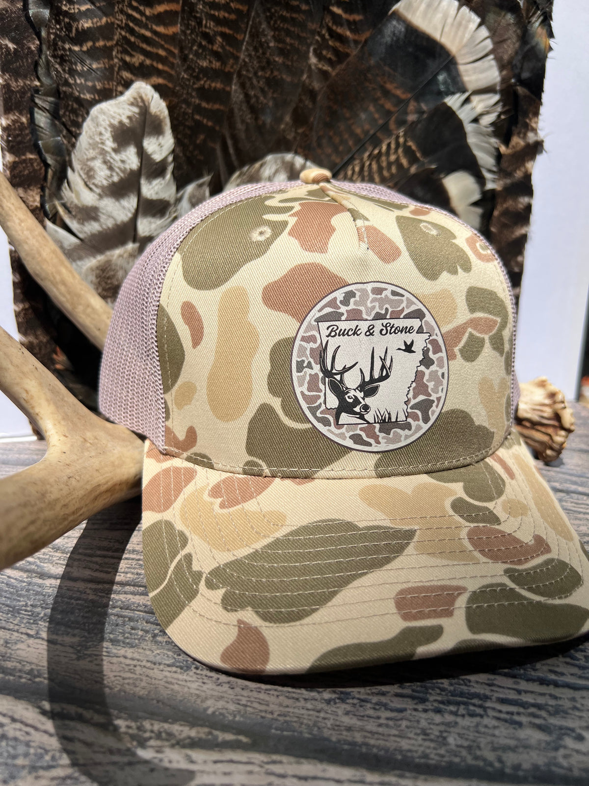 A camouflage trucker hat with a circular 'Back & Stone' logo patch on the front, styled with deer antlers and a feathered headband on a rustic wooden surface.