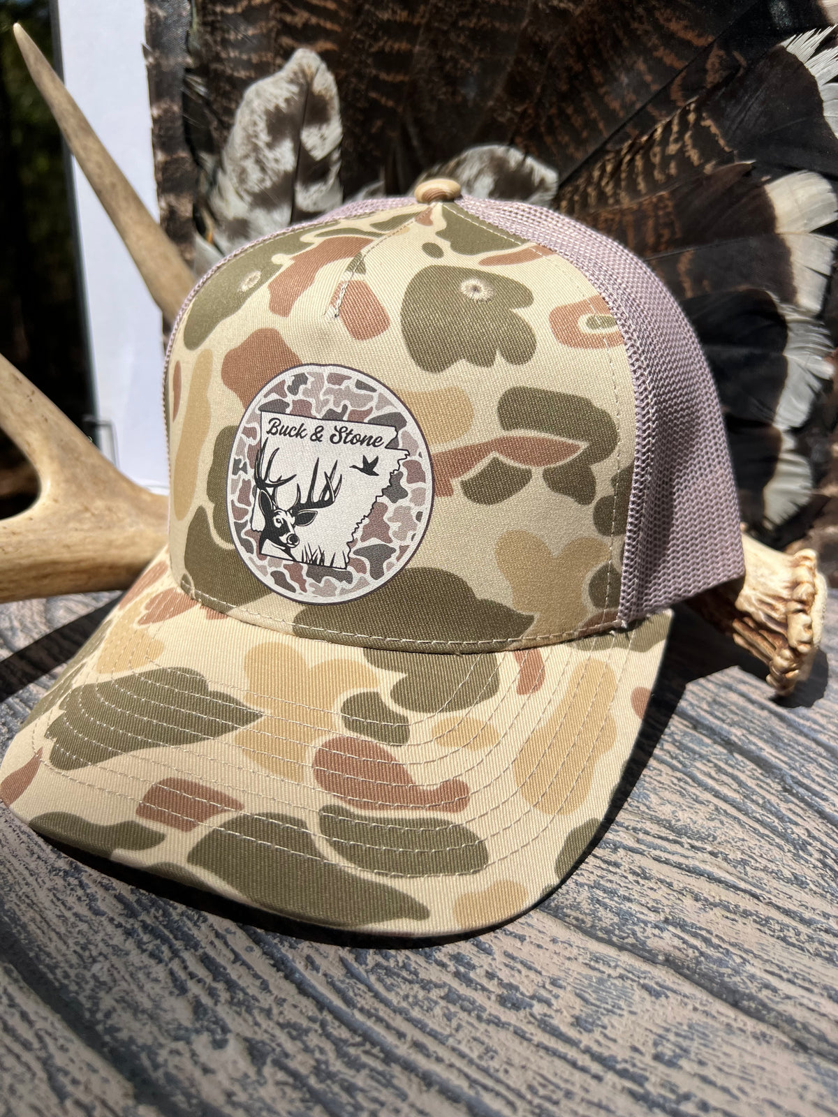 A camouflage trucker cap with a round 'Buck & Stone' logo patch on the front, resting on a rustic wooden surface with deer antlers and feathers in the background.
