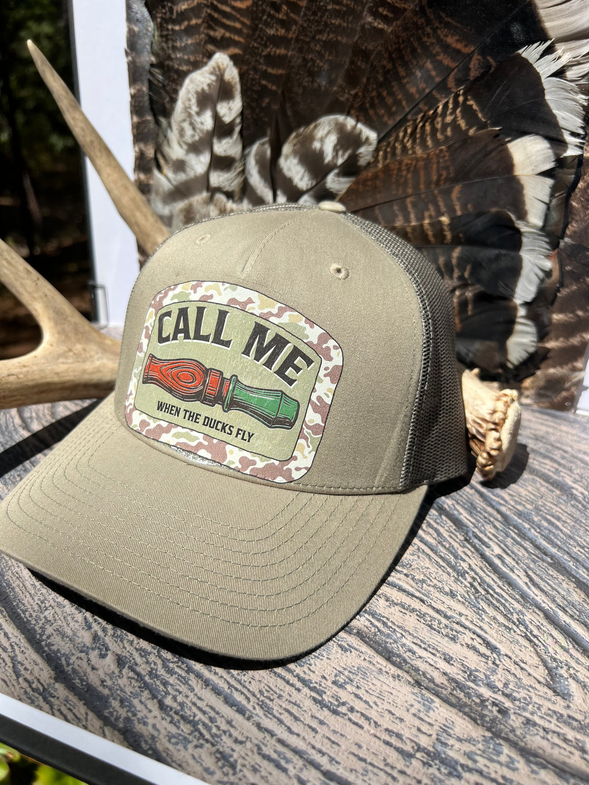 An olive green trucker hat with a camouflage patch on the front that reads 'CALL ME WHEN THE BUCKS FLY' with a red and green bowie knife graphic. The hat is styled on a rustic wooden surface with deer antlers and a large turkey feather in the background.