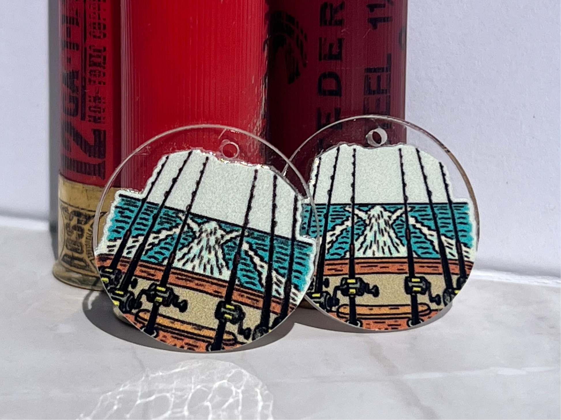 Round earrings with fishing reel design on a white surface with red and gold cylindrical objects in the background.