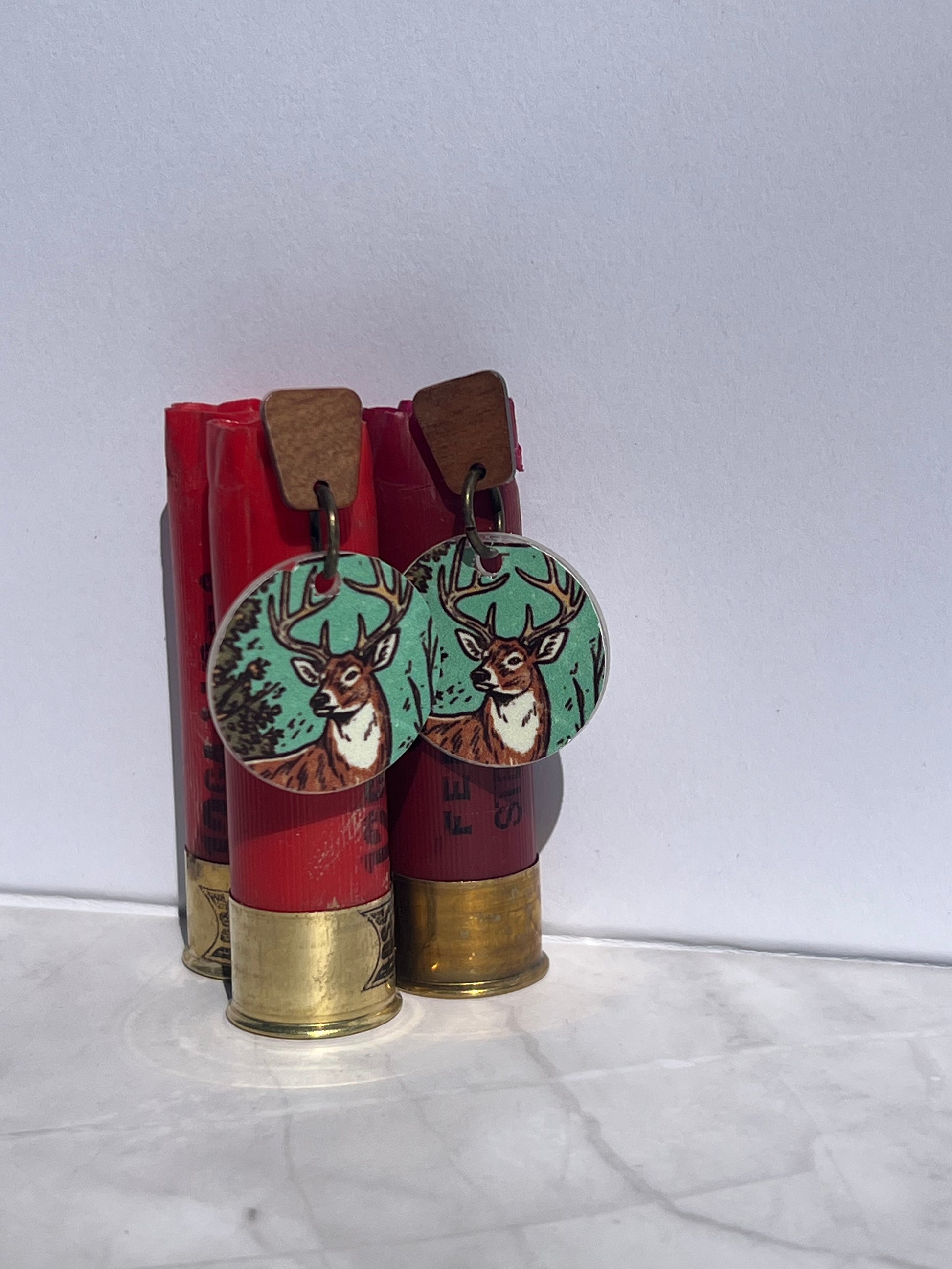 Deer-themed earrings with wood post displayed on red shotgun shells against a white background